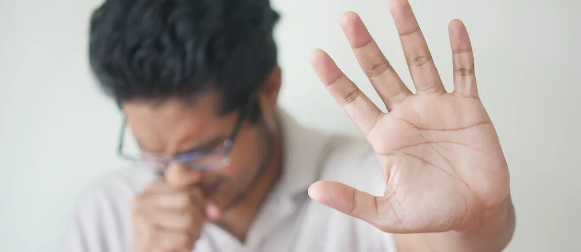 Ein Mann hustet in die eine Hand und hält die andere Hand schützend vor sich.