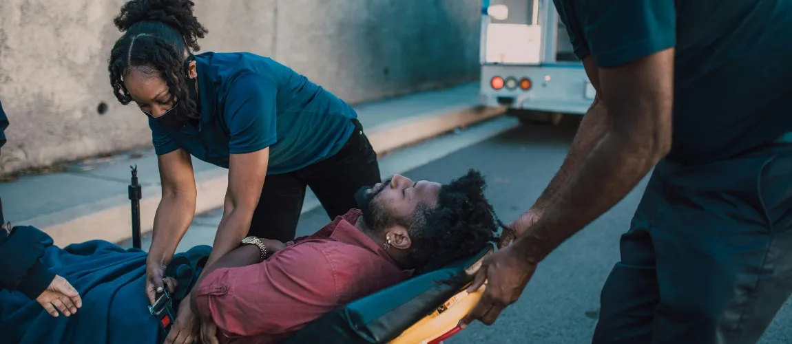 Ein Mann liegt auf einer Transportliege, zwei Pflegefachkräfte kümmern sich um ihn. 