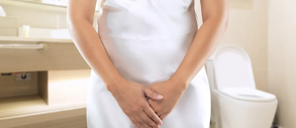 Eine Person hält die Hände vor dem Unterleib verschränkt, im Hintergrund ist eine Toilette zu sehen.