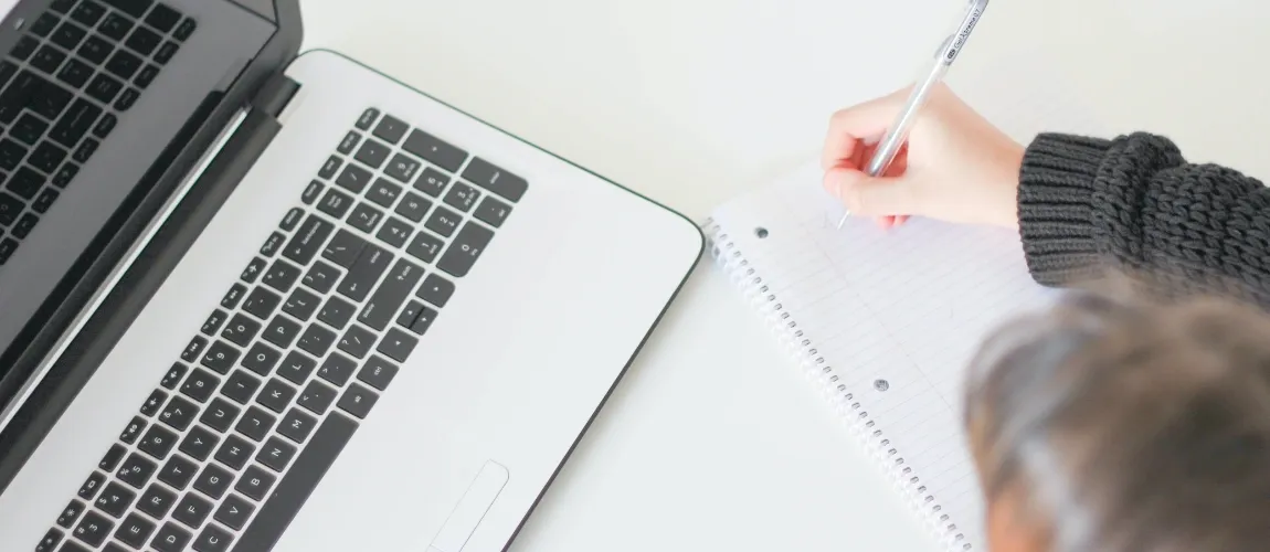 Eine Person macht Notizen in einem Schreibblock, vor ihr steht ein Laptop. 