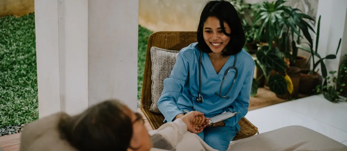 Eine Pflegekraft sitzt vor dem Bett einer pflegebedürftigen Person und hält ihre Hand.