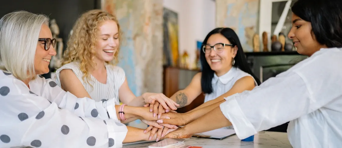 Vier Frauen sitzen an einem Tisch und legen die Hände aufeinander.